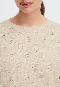 Beige strikket genser med et teksturert mønster, med rund hals og delikate sømdetaljer gjennom hele.