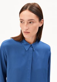 Vrouw met steil bruin haar die een blauwe blouse met kraag draagt, kijkt naar links tegen een effen witte achtergrond.