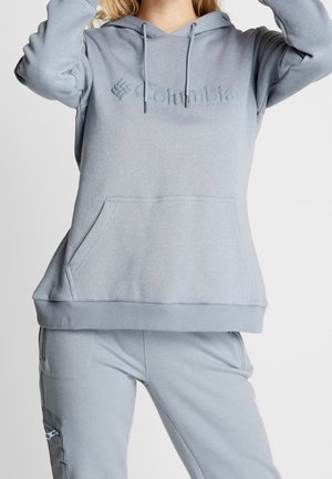 Mujer que lleva una sudadera con capucha Columbia de color gris claro y pantalones a juego con una cremallera en el bolsillo lateral sobre un fondo liso.