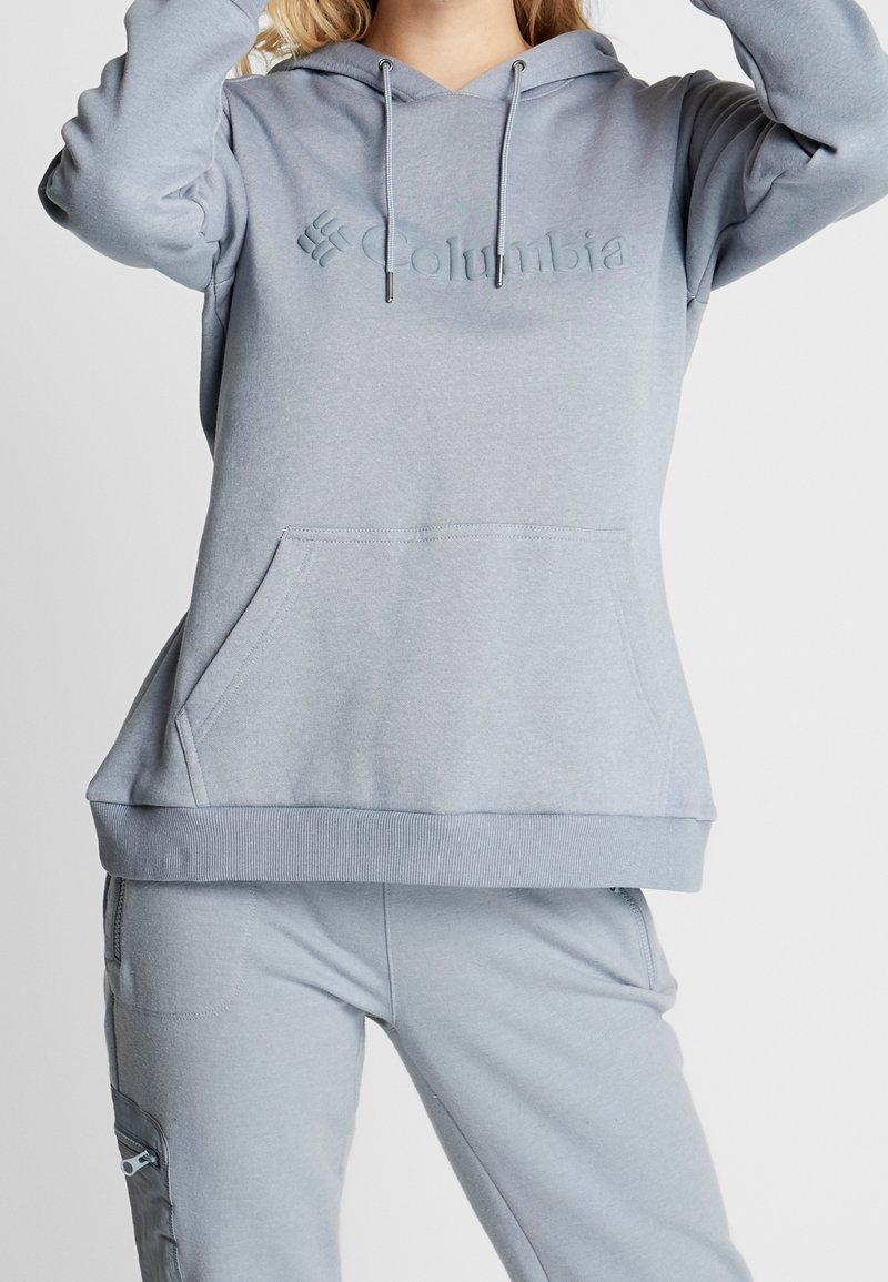 Femme portant un sweat à capuche Columbia gris clair et un pantalon assorti avec une fermeture éclair sur la poche latérale devant un fond uni.