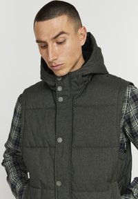 Dark green, hooded vest with a quilted texture. Features silver snap buttons and a layered plaid shirt underneath with dark and light patterns.
