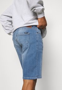 Pantalones cortos de mezclilla en azul claro, con un corte recto, diseño de cinco bolsillos y una etiqueta de cuero marrón en la cinturilla. Textura suave.