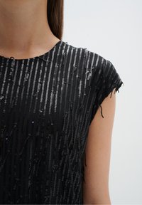 Černý top bez rukávů s vertikálními flitry různých velikostí a struktur, vytvářející vrstvený, třpytivý efekt na jednolitém pozadí.