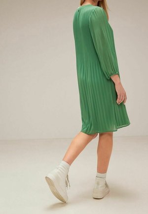 Zielona, plisowana sukienka z długimi rękawami, z okrągłym dekoltem i guzikami z tyłu; zestawiona z białymi, wysokimi trampkami.