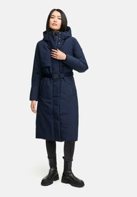 Lungo cappotto puffo blu navy con cappuccio rimovibile, vita con cintura, tasche con zip e texture liscia. Indossato con jeans scuri e stivali.