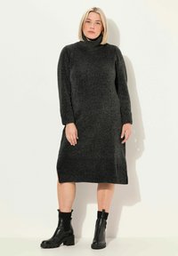 Robe en tricot noir à col roulé avec manches longues et fentes sur les côtés, fabriquée dans un tissu texturé. Portée avec des bottines noires.
