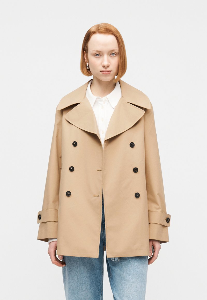 Γυναίκα με ίσια κόκκινα μαλλιά φοράει μπεζ διπλόμπουτο παλτό, λευκό πουκάμισο με γιακά και ανοιχτό μπλε τζιν, στέκεται μπροστά σε λευκό φόντο.