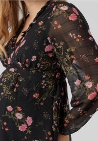 Mustakukkainen mekko, jossa on läpinäkyvät pitkät hihat ja V-pääntie. Korostettu vaaleanpunaisilla ja keltaisilla kukilla, teksturoidulla kankaalla ja vyötäröltä korostavalla mitoituksella.