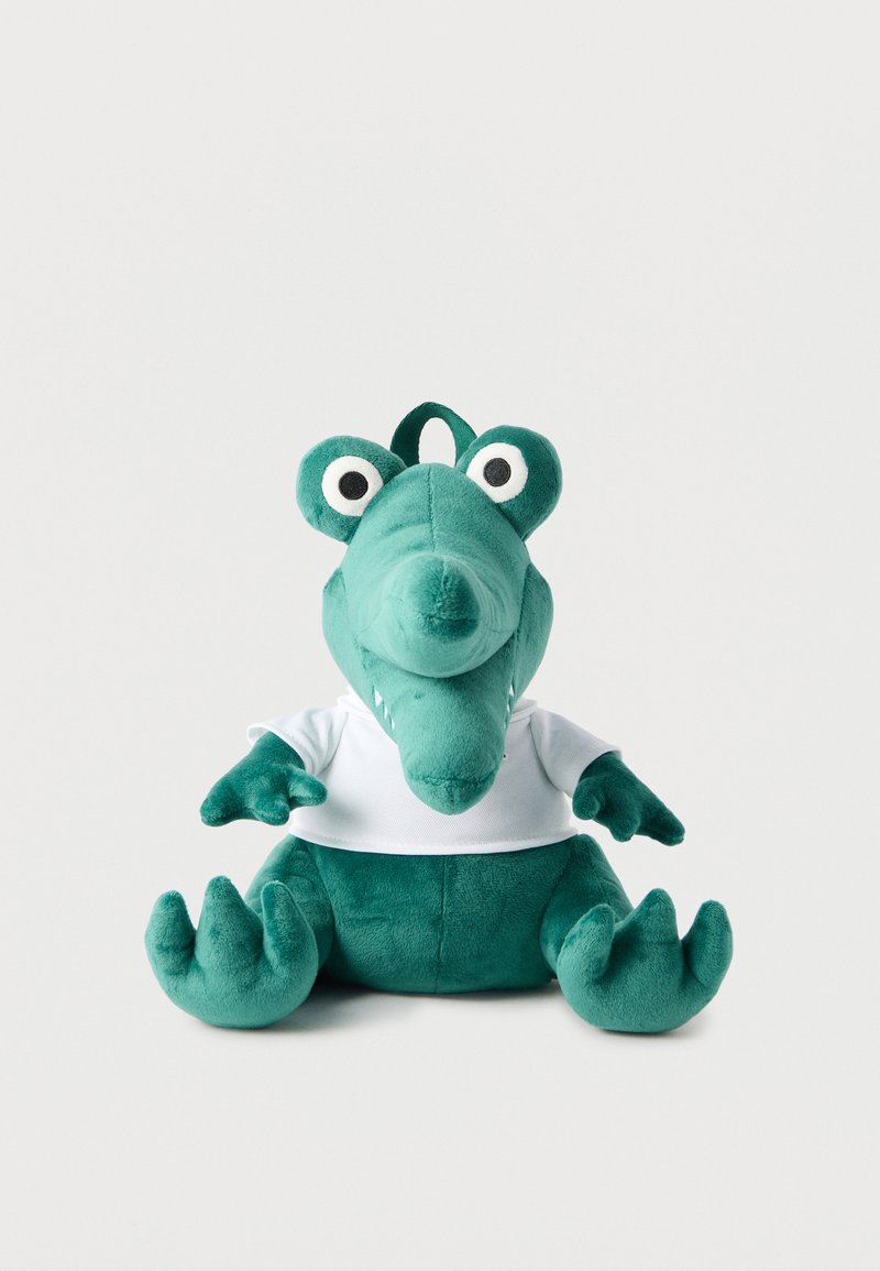 Giocattolo di coccodrillo di peluche verde che indossa una maglietta bianca, seduto su uno sfondo bianco uniforme con un anello di tessuto sulla testa.