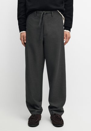 Mann trägt lockere dunkelgraue Hose mit Kordelzug am Bund, kombiniert mit dunkelbraunen Schnürschuhen und einem schwarzen Pullover.