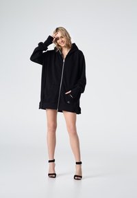 Czarna bluza z kapturem na zamek, wykonana z miękkiego materiału, z dużym kapturem, kieszeniami z przodu i ściągaczami na rękawach. Noszona na szorty i szpilki.