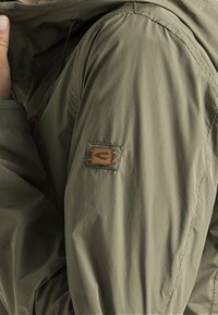 Veste en nylon vert olive avec une surface texturée ; comporte un patch signature sur la manche et une fermeture de cou ajustable.
