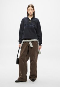 Μπλε σκούρο φούτερ με ψηλό γιακά και κεντημένο λογότυπο, σε συνδυασμό με άνετο, καφέ παντελόνι από βελούδο κορντούρο και ανοιχτό γκρι πουλόβερ δεμένο στη μέση.