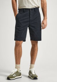 Pantalones cortos de algodón azul marino con bolsillos laterales, cierre de botón y un corte relajado, combinados con zapatillas verde oliva y calcetines beige.