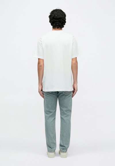 Biała bawełniana koszulka z krótkim rękawem, widziana od tyłu, zestawiona z jasnoniebieskimi dżinsami i sneakersami w neutralnych tonacjach.