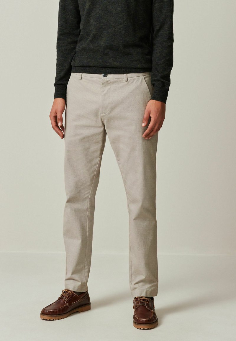 Pantalones beige con un acabado texturizado, que cuentan con bolsillos frontales, trabas para cinturón y un cierre de botón, combinados con zapatos marrones con cordones.