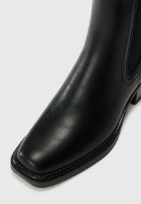 Bottine en cuir noir avec un bout carré, texture lisse et panneaux latéraux en elastique. Présente un petit talon solide et des détails de couture discrets.