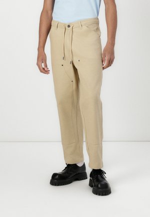 Beige trousers with a drawstring waist, side pockets, and contrasting dark hardware. Paired with black shoes featuring a thick sole.