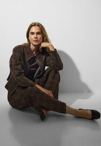 Femme aux longs cheveux blonds portant un costume marron à carreaux et des ballerines noires, assise par terre, le menton appuyé sur la main contre un mur blanc.