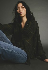 Blouse noire, légère et transparente avec un motif à carreaux et une coupe décontractée, associée à un jean en denim bleu clair. Texture lisse visible.
