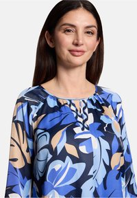 Blouse fleurie en marine, bleu et beige ; tissu léger ; encolure ronde avec un détail œillet ; manches trois-quarts.