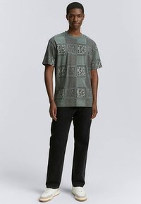 Zielony t-shirt we wzory z krótkimi rękawami i okrągłym dekoltem, zestawiony z czarnymi jeansami i białymi trampkami z jasnobrązowymi akcentami.