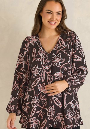 Vrouw draagt een donker lange mouwen V-hals blouse met roze en witte bloemenpatronen, glimlacht zachtjes, hand rust op haar buik.