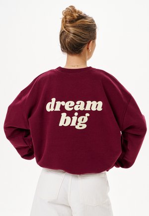 Vrouw met haar in een knot, die een bordeauxrode trui draagt met de tekst "dream big" op de rug en witte broek, met haar rug naar de camera.