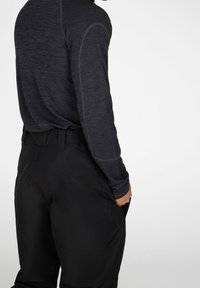Homme portant un pantalon noir et une chemise sombre à manches longues, vu de dos et de côté avec une main dans une poche.