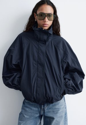 Femme portant une veste oversize bleu marine foncé avec col haut et poignets élastiques, jean bleu clair, et grandes lunettes de soleil teintées, debout contre un mur blanc.