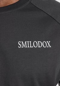 Musta t-paita, joka on valmistettu pehmeästä ja joustavasta kankaasta, jossa brändin nimi "SMILODOX" on kirjailtuna valkoisena rintakappaleessa. Pyöreä pääntie.
