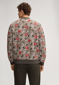 Giacca bomber trapuntata in verde oliva con motivi bianchi svolazzanti e labbra rosse. Maniche e fondo a costine, con una finitura testurizzata.