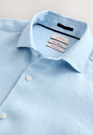 Camisa de lino azul claro con un corte regular, que presenta un cuello abotonado y tres etiquetas internas que muestran el nombre de la marca y detalles del tejido.
