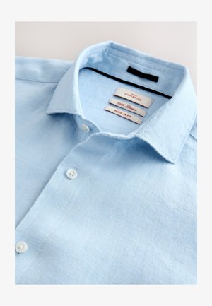 Camisa de lino azul claro con un corte regular, que presenta un cuello abotonado y tres etiquetas internas que muestran el nombre de la marca y detalles del tejido.
