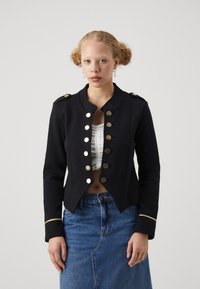 Veste noire cropped avec des boutons dorés, design structuré et accents dorés sur les manches, associée à un haut blanc à volants et une jupe en denim.