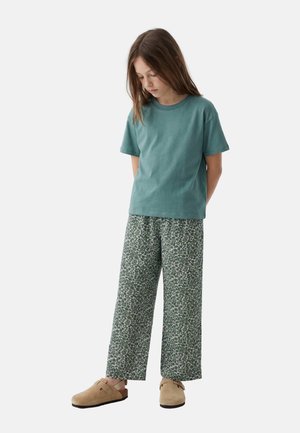 Jeune fille aux longs cheveux portant un t-shirt turquoise à manches courtes, un pantalon vert à motifs à jambes larges et des chaussures beiges à enfiler, debout les mains dans le dos.