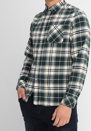 Mann trägt grün-weiß-gelb kariertes Hemd mit Brusttasche, Hände in dunklen Hosentaschen, steht vor einfachem Hintergrund.