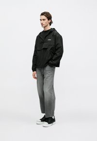 Νέος άνδρας που φοράει μαύρο αδιάβροχο με κουκούλα, γκρι τζιν και μαύρα αθλητικά παπούτσια, στέκεται μπροστά σε απλό λευκό φόντο.