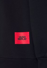 Svart tyg med en vävd textur och en röd rektangulär etikett. Etiketten har texten "HUGO REPLAY" i svarta bokstäver.