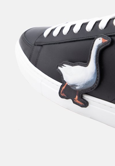 Baskets noires avec semelle et lacets blancs, ornées d'un patch oie blanche sur le côté près de la zone des orteils.