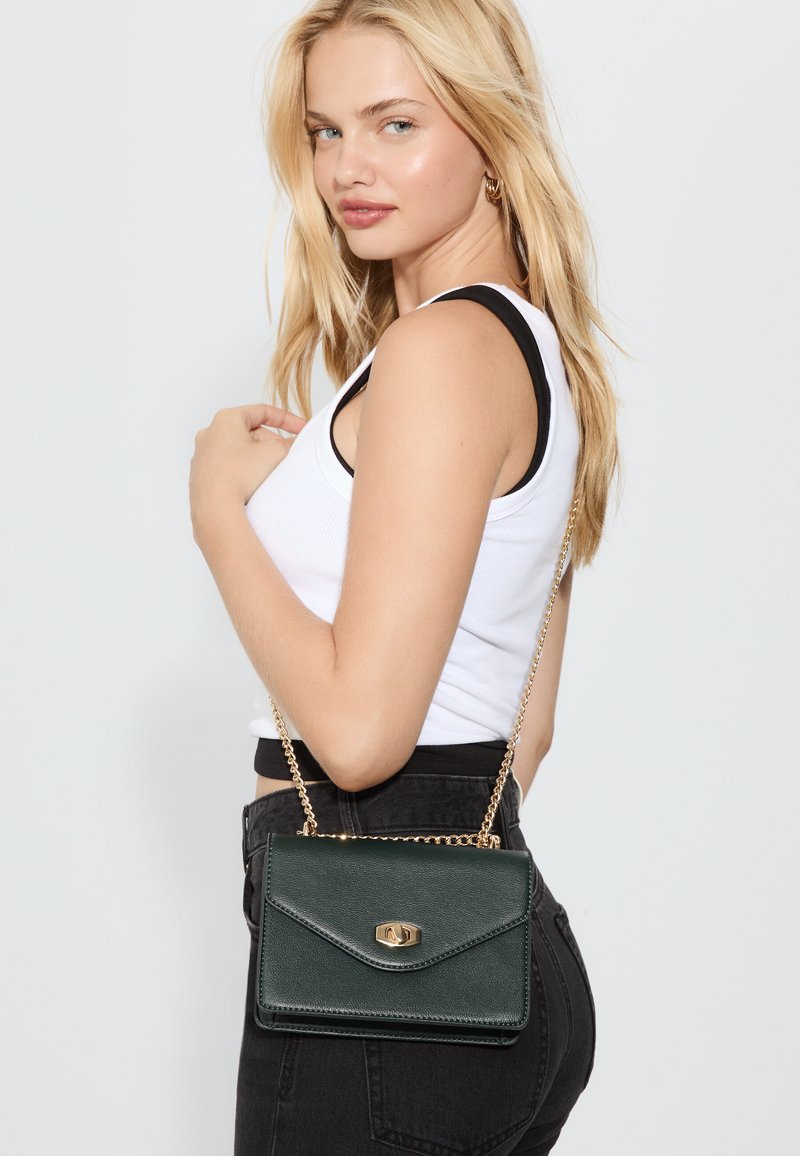 Grüne Umhängetasche aus Kunstleder mit goldfarbenem Verschluss und Kettengurt, ausgestattet mit einem Kuvert-Design und strukturierter Oberfläche.