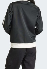 Černá sportovní bunda s bílými akcenty, vyrobená z hladké, lehké látky; má klasický límec a žebrované manžety a lem.