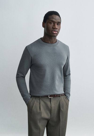 Homme debout les mains dans les poches, portant une chemise grise à manches longues et un pantalon plissé marron, sur un fond clair uni.