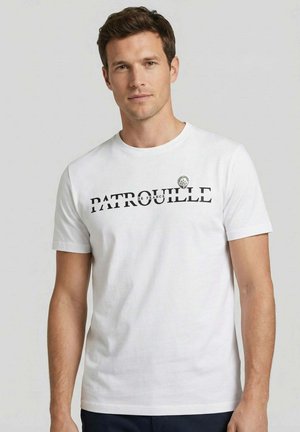 Homme portant une chemise blanche à manches courtes avec le texte « PATROUILLE DE FRANCE » et un petit emblème sur la poitrine, debout devant un fond gris clair.