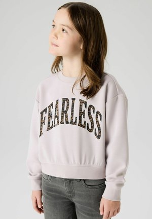 Jeune fille aux cheveux châtain clair portant un sweat-shirt léger avec le texte "FEARLESS" en imprimé léopard, regardant vers le haut à gauche.