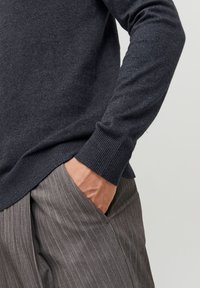 Personne portant un pull gris foncé à manches longues avec une main dans la poche d’un pantalon gris à fines rayures sur un fond uni.
