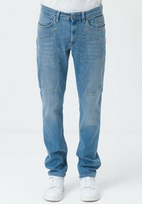 Jeans in denim azzurro chiaro con una vestibilità rilassata, caratterizzati da ampie tasche frontali e un leggero sbiadimento. Abbinati a scarpe da ginnastica bianche su uno sfondo neutro.