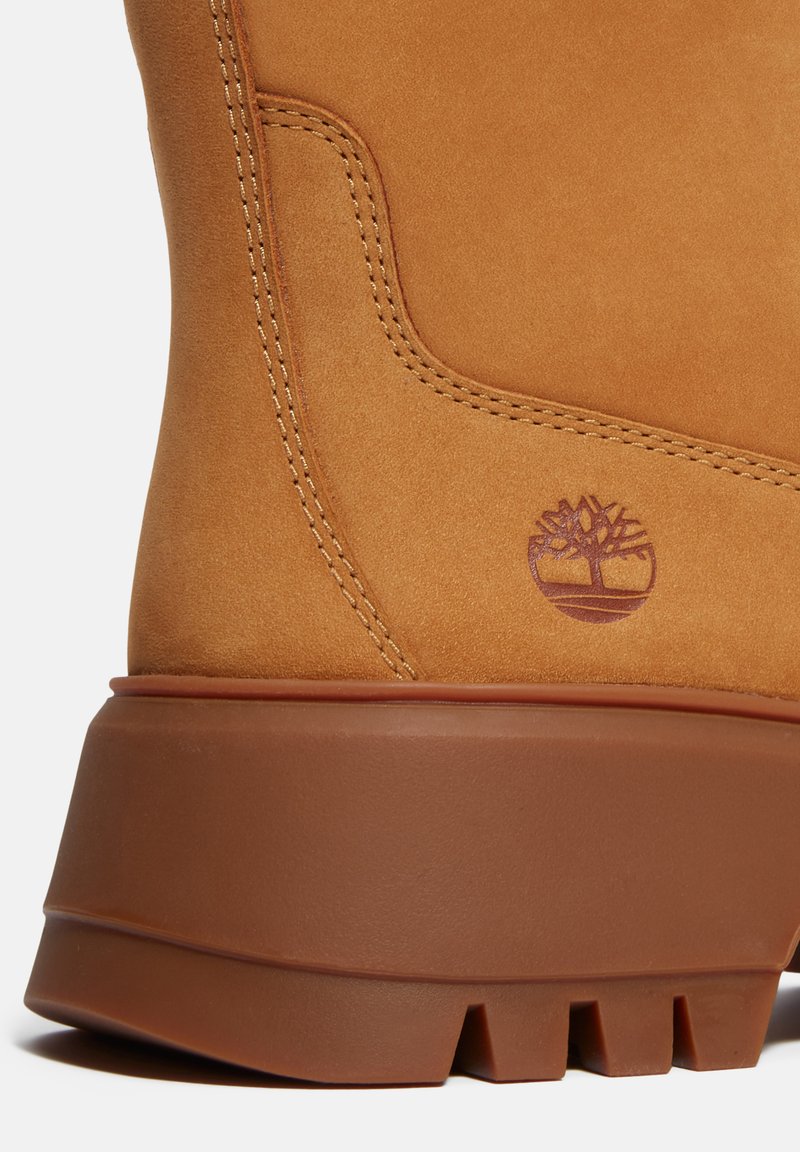 Stivale in suede marrone chiaro con suola in gomma marrone chunky, caratterizzato da cuciture gialle e un piccolo logo a forma di albero viola sul lato.