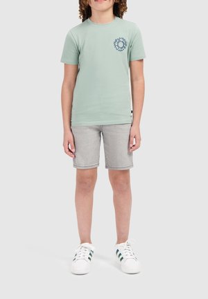 Mintgroene t-shirt met korte mouwen en een cirkelvormige blauwe grafiek, gecombineerd met lichtgrijze shorts en witte sneakers met groene strepen.