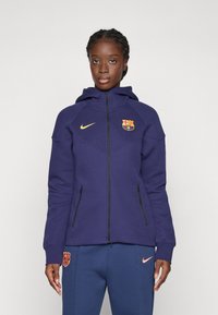 Lila Zip-Hoodie mit Kapuze, versehen mit dem Barcelona-Wappen und dem Nike-Logo in Gold. Strukturierter Stoff und zwei mit Reißverschluss versehenen Seitentaschen.